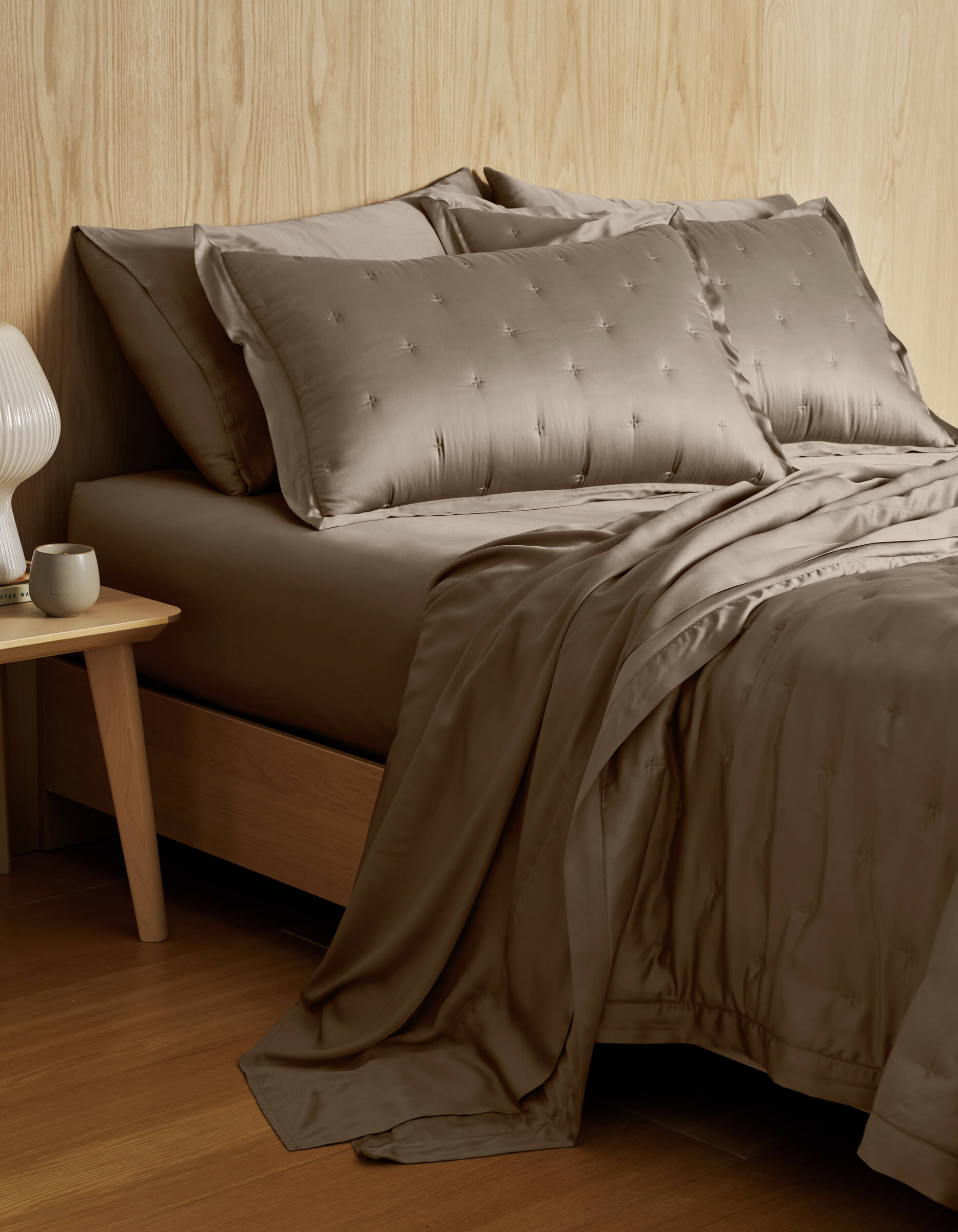 A neatly made bed featuring the Cozy Earth Bamboo Quilt and matching pillows in taupe sits beside a wooden nightstand with a white cup and modern lamp, set against a light wood-paneled wall.