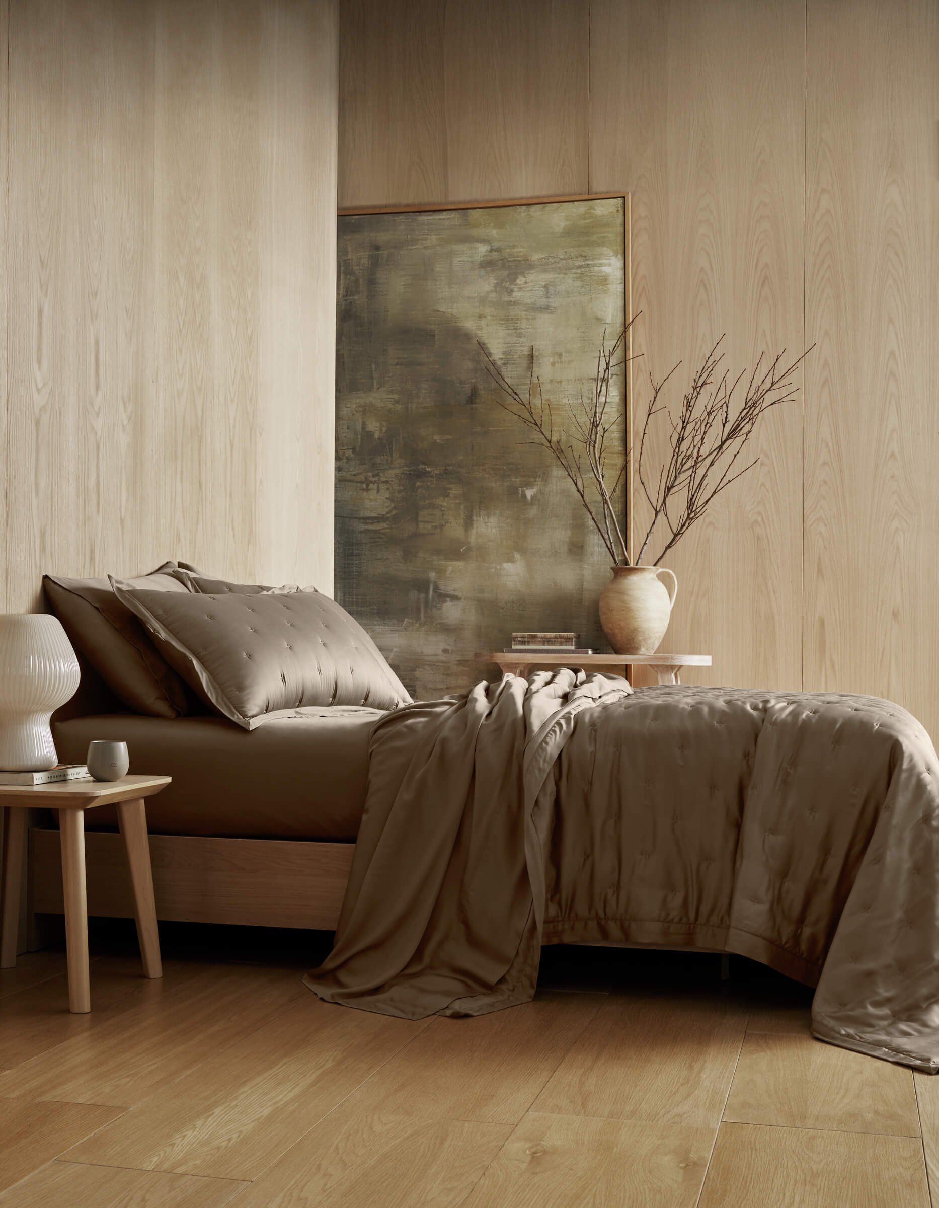 A modern bedroom features the Cozy Earth Bamboo Quilt Bundle with taupe bedding and matching pillows. Decorative touches include a beige vase with branches, a white lamp, and a small side table against wooden paneling and an abstract painting.