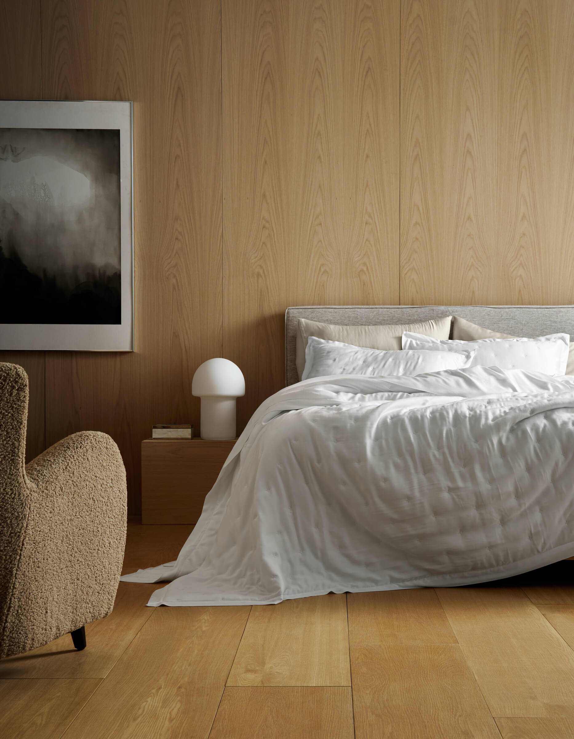 A modern bedroom features light wood panels, a bed made with Cozy Earth’s Bamboo Quilt, a beige armchair, a wooden nightstand with a white lamp, and abstract black-and-white artwork on the wall.