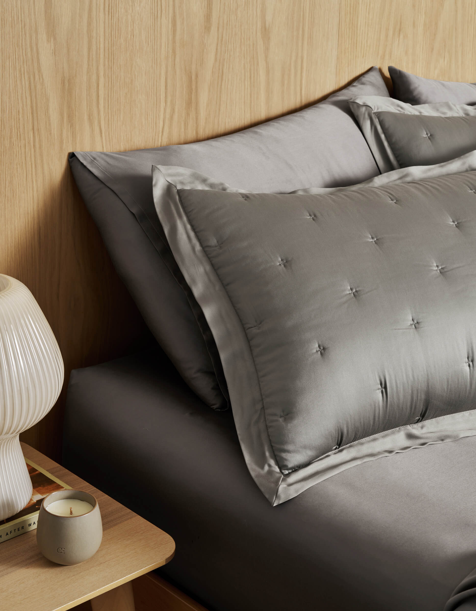 A neatly made bed with Cozy Earth's Bamboo Quilted Shams and tufted pillows sits beside a wooden nightstand with a ribbed white lamp, a candle, and books, set against a light wood-paneled wall.|Color:Charcoal