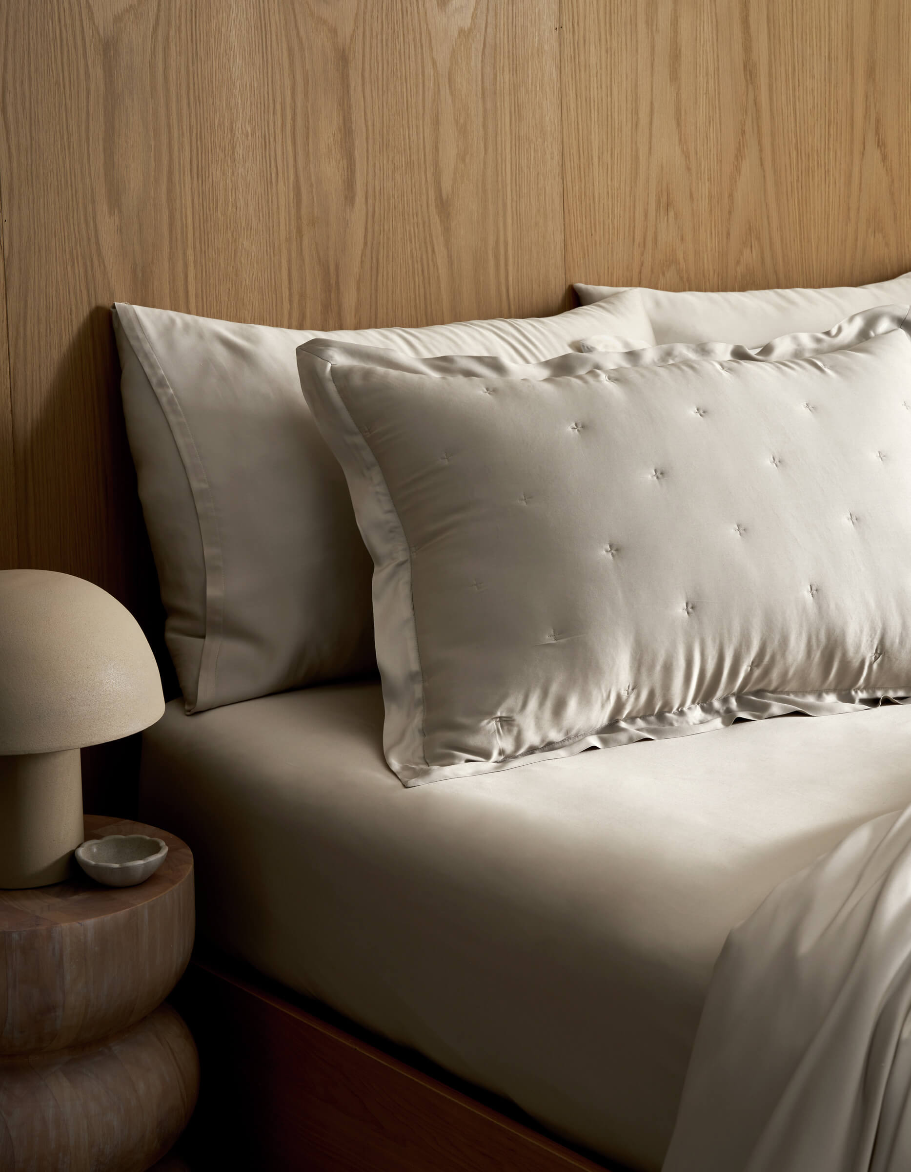 A neatly made bed features Cozy Earth's Bamboo Quilted Shams in light tones, set against a wooden headboard. A round bedside table holds a dome-shaped lamp and a small dish.