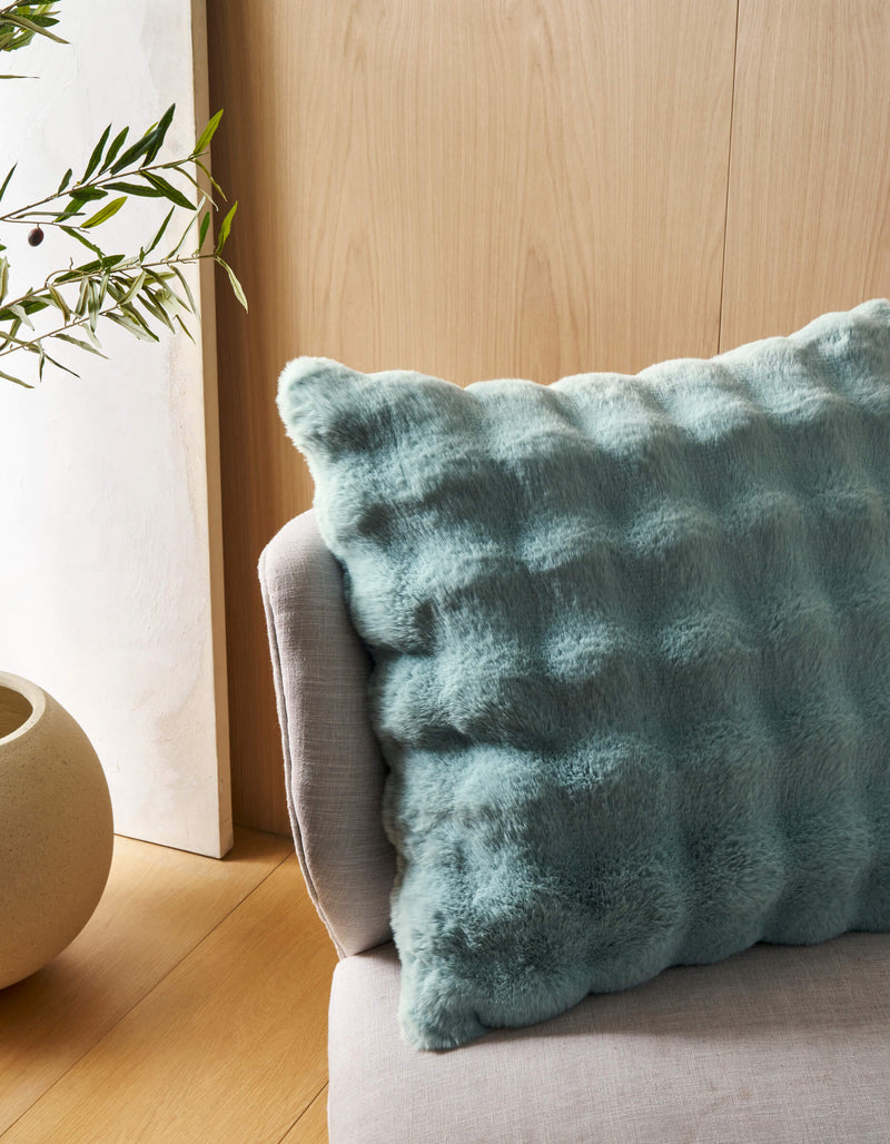 The Cozy Earth Bubble Cuddle Pillow in soft textured light blue sits on a light gray chair beside a beige pot with a leafy green plant, set against a light wood wall and floor.