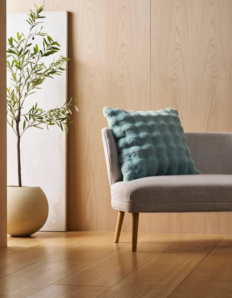 A light gray chair with wooden legs holds a Cozy Earth Bubble Cuddle Pillow in textured blue. Next to it is a potted plant in a beige planter, set before a wooden wall with a minimalist white art piece behind the plant.