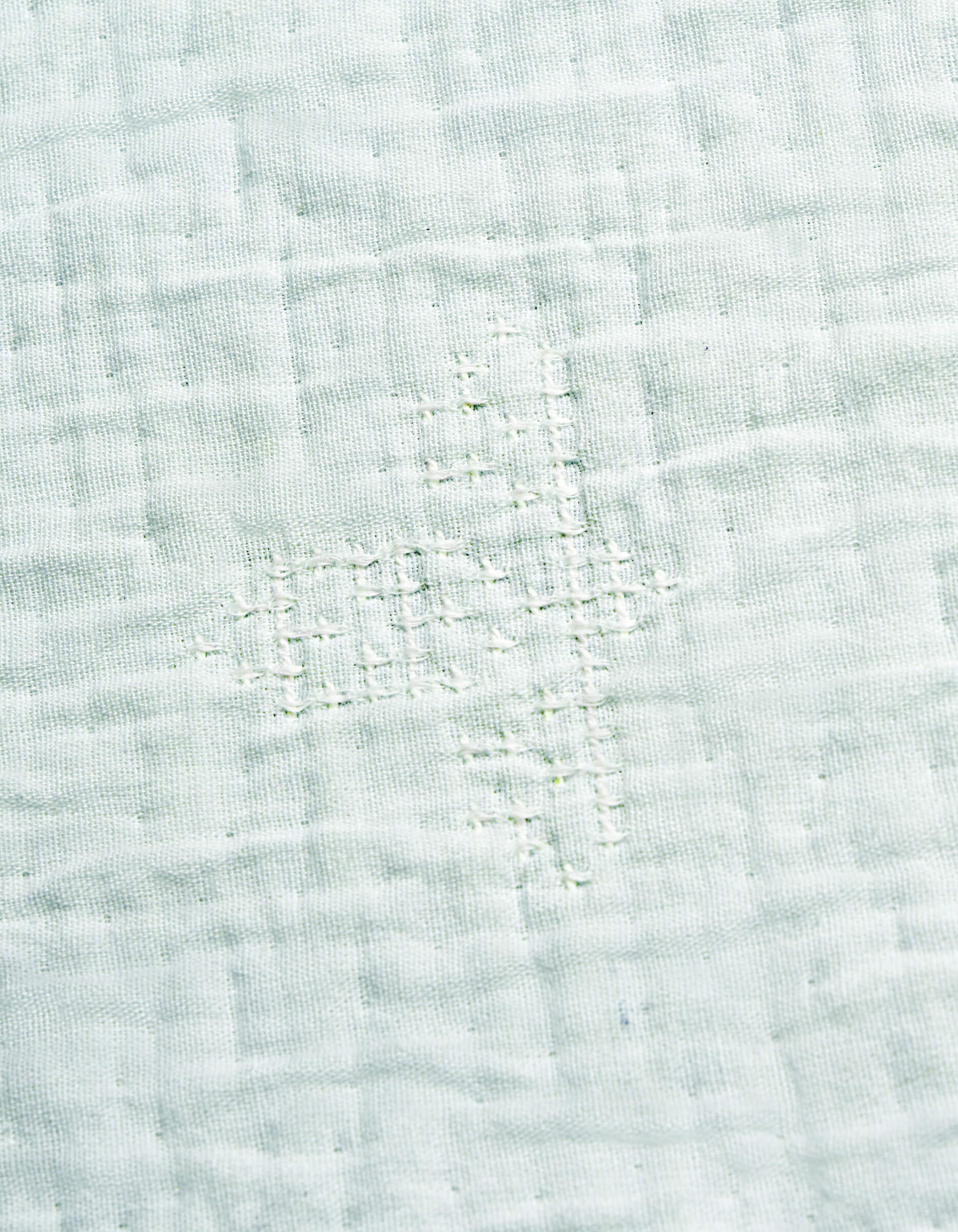 A close-up of the Cozy Earth Little Bee Matelasse Baby Blanket shows its white fabric with a grid-like, hand-stitched repair in matching thread, highlighting the textured and slightly uneven surface.