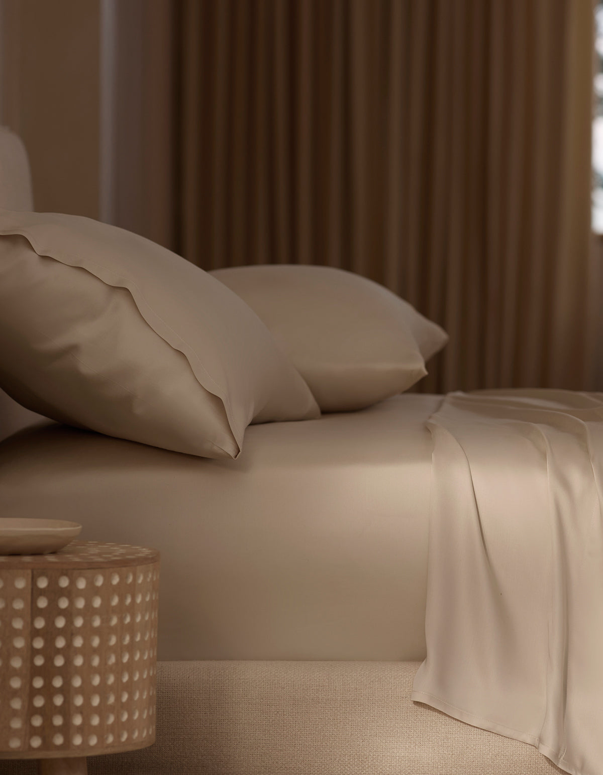 A neatly made bed features Cozy Earth’s Bamboo Sheet Set in beige with matching pillows, beside a round wooden nightstand with a dotted design. Soft, warm light through curtains enhances the cozy atmosphere. |Color:Driftwood