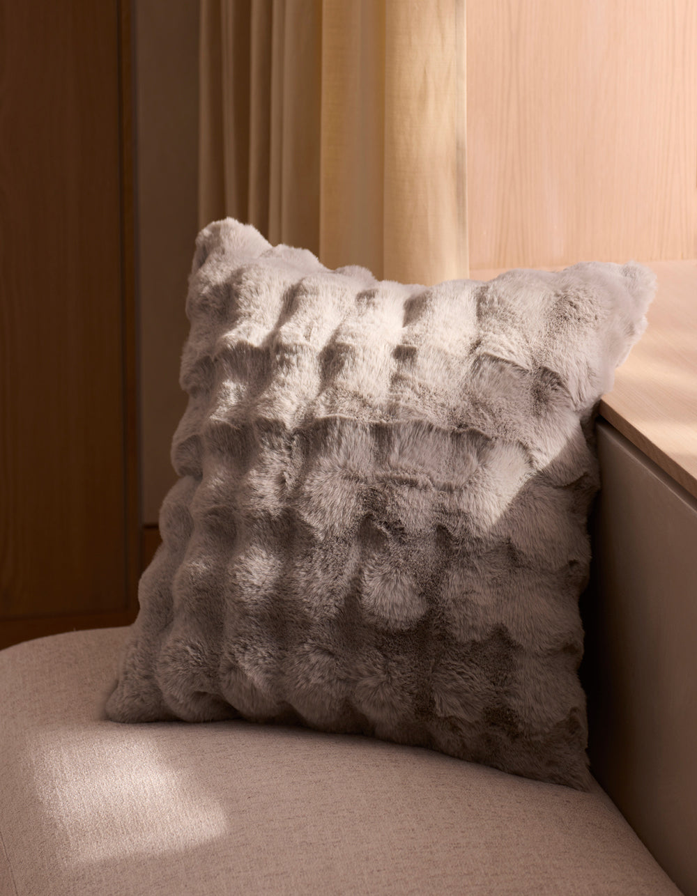 The Bubble Cuddle Pillow by Cozy Earth, in light gray textured faux fur, sits on a beige bench beside a wooden wall and cream curtains, bathed in soft natural light.