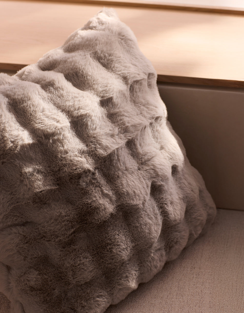 The Cozy Earth Bubble Cuddle Pillow, featuring soft, textured light grey faux fur, rests on a beige surface as natural light casts gentle shadows.