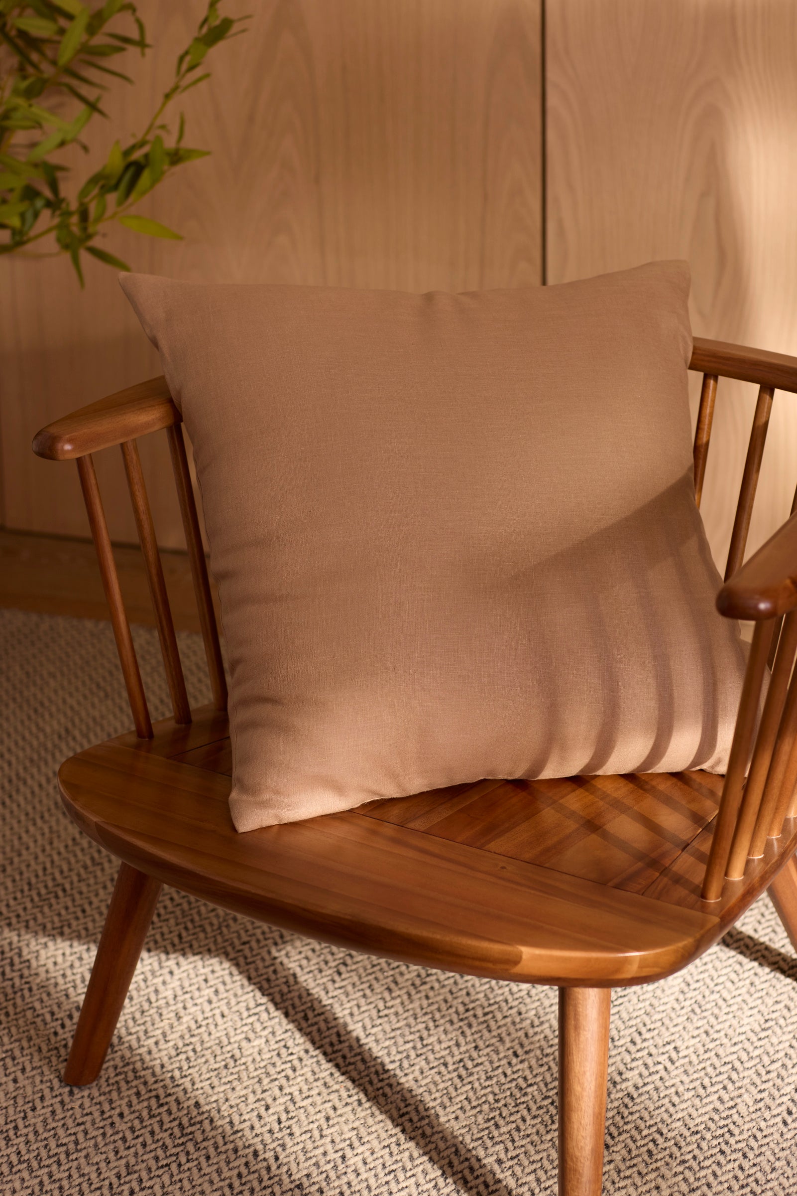 The Cozy Earth Linen Throw Pillow rests on a wooden spindle-back chair, placed on a textured rug near a wooden wall and green leafy plant, with warm sunlight casting soft shadows across the scene. |Color:Maple|Size:24" x 24"