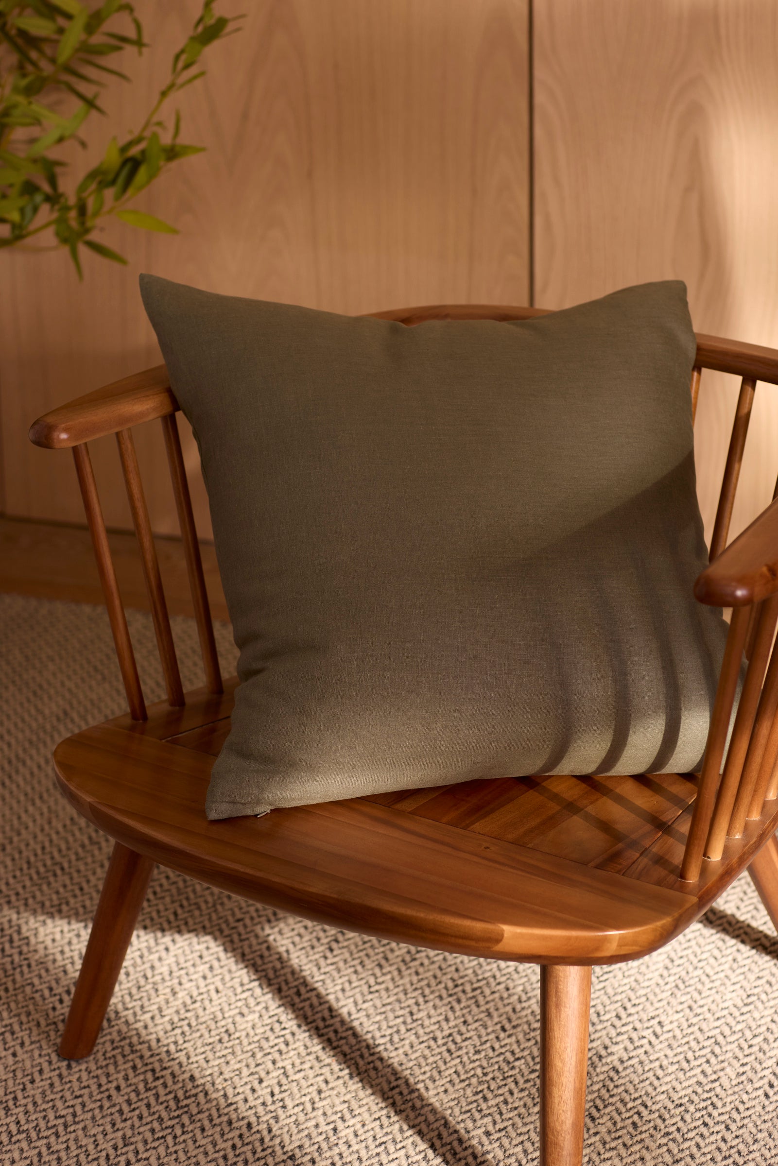 A Cozy Earth Linen Throw Pillow rests on a wooden chair atop a textured rug. Sunlight casts soft shadows across the chair and pillow, while a leafy plant is partially visible in the background.