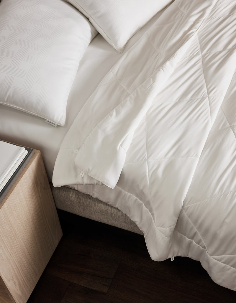 A neatly made bed features white pillows and the Cozy Earth Bamboo Viscose Comforter, partially folded back. Beside it, a light wood nightstand with a book rests on dark wood floors.