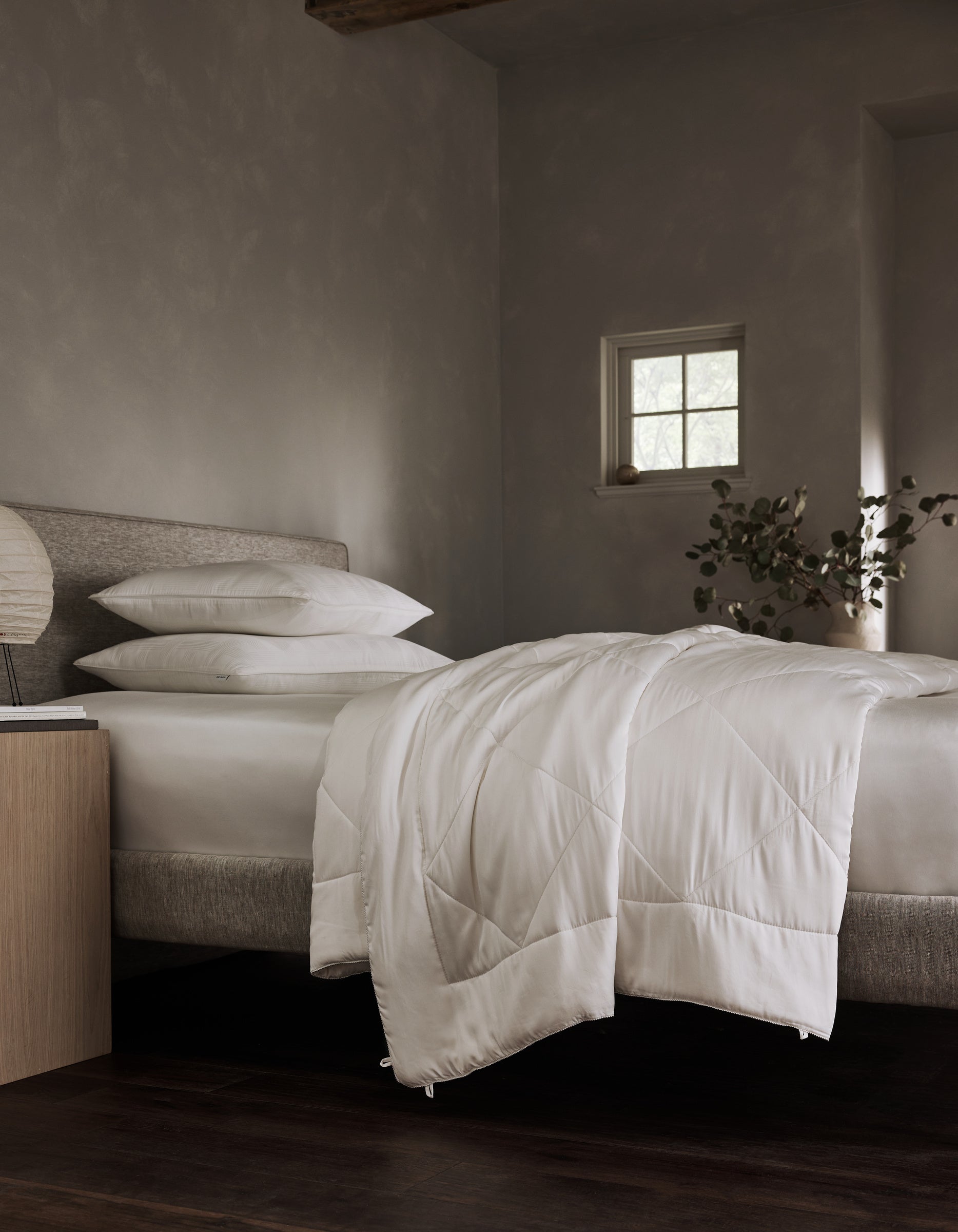 A bed dressed in Cozy Earth's Bamboo Viscose Comforter with white pillows anchors a minimalist bedroom featuring gray walls, a small window, a nightstand with lamp, and a vase of greenery.