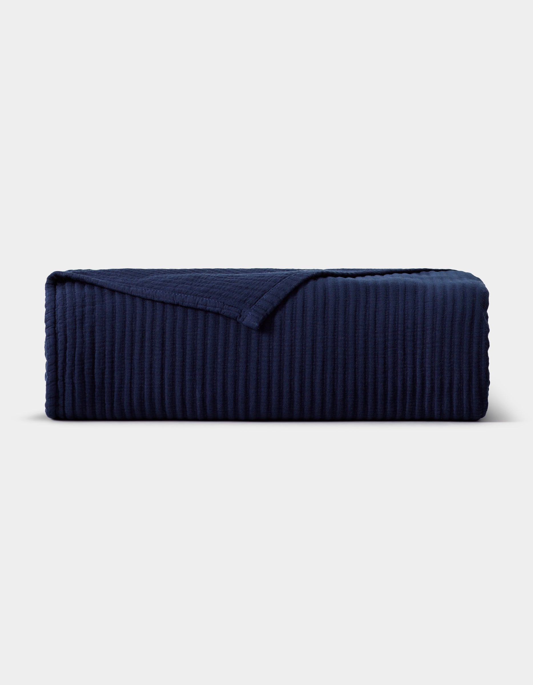 The Cozy Earth Bamboo Coverlet, in dark navy blue with a ribbed texture and one corner turned down, is displayed neatly against a plain light gray background.