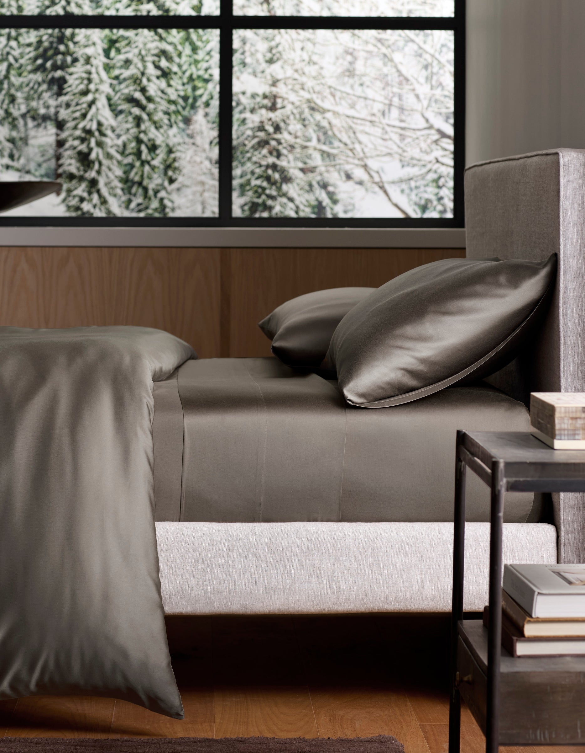 A neatly made bed with Cozy Earth’s Bamboo Sheet Set in dark gray rests by a modern nightstand. Behind the bed, a large window reveals a snowy forest with pine trees. 