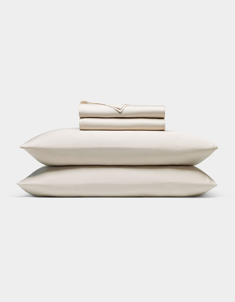 A neatly folded Cozy Earth Bamboo Sheet Set—cream-colored, including a fitted sheet, flat sheet, and two pillowcases—rests atop two matching pillows against a plain white background.