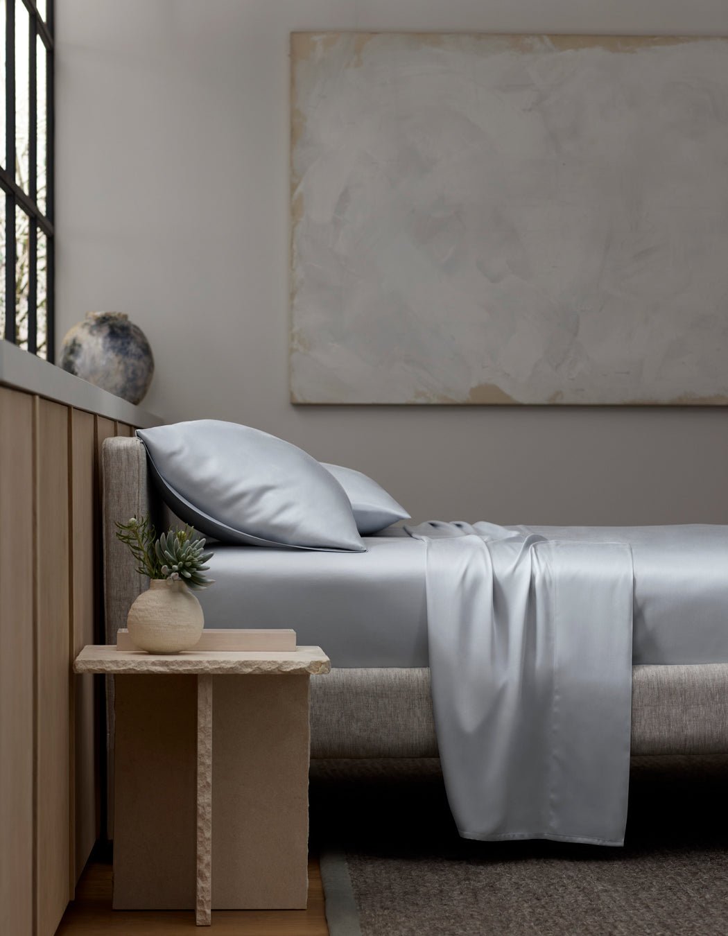 A minimalist bedroom with a light gray upholstered bed, Cozy Earth Bamboo Sheet Set in silver-gray, two pillows, a beige nightstand with a small potted plant, a large abstract painting, and a window letting in natural light.|Color:Shore