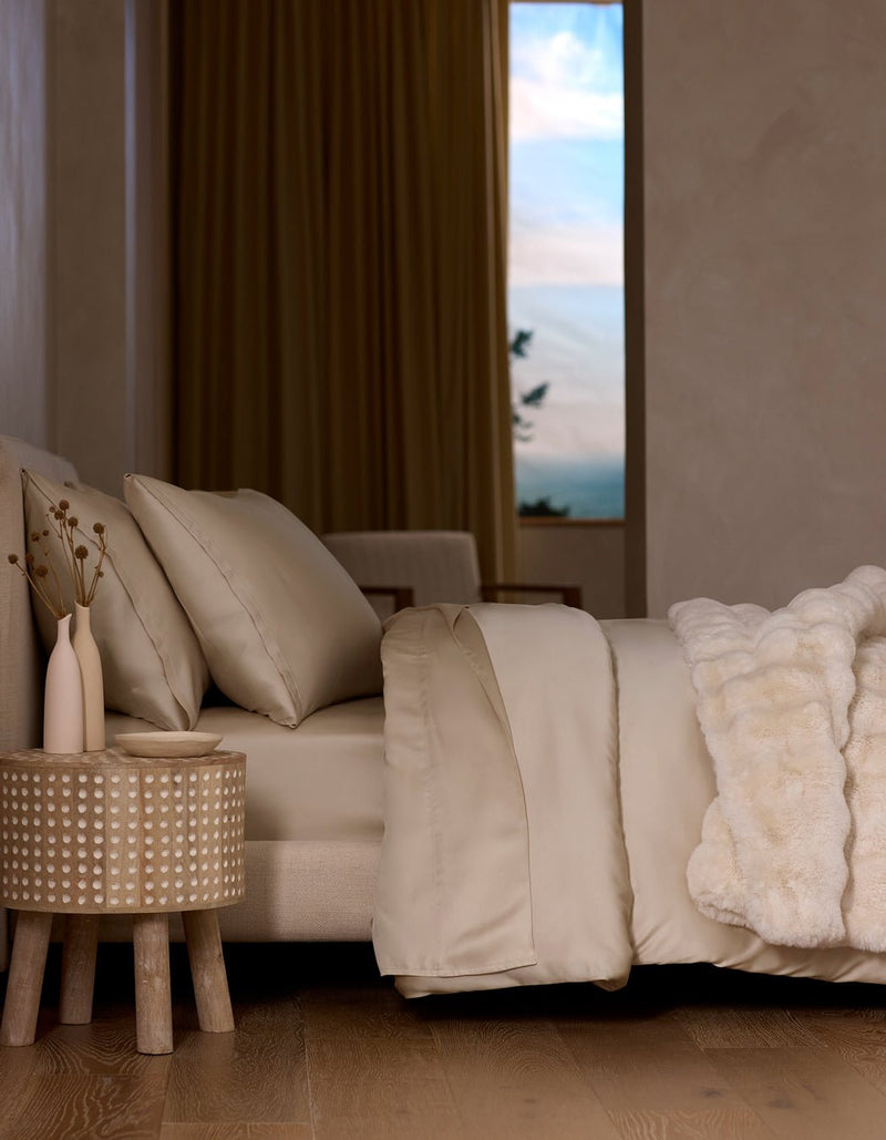 A neatly made bed features the Cozy Earth Bamboo Duvet Cover in beige and a white faux fur blanket, beside a textured wooden nightstand with two vases. Large curtained windows reveal a scenic sky.
