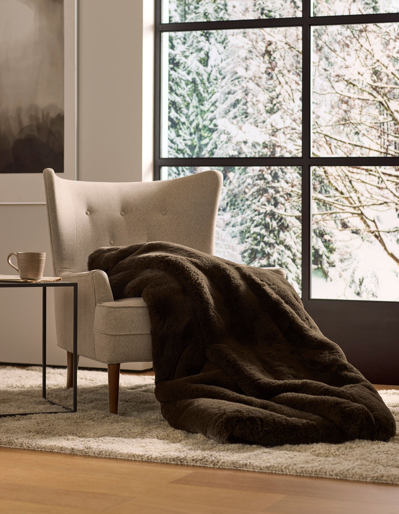 A cozy beige armchair with a Cozy Earth Cuddle Blanket draped over it sits next to a small table with a mug, overlooking snowy trees through large windows.|Color:Espresso
