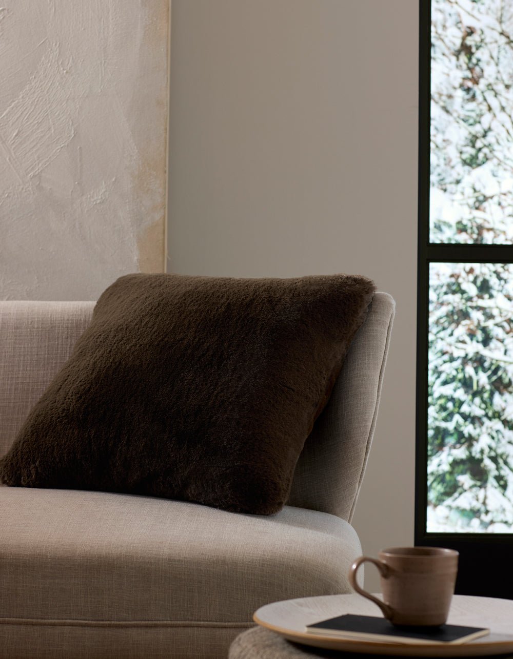 A beige sofa with the Cozy Earth Cuddle Pillow in dark brown sits by a window overlooking snow, while a ceramic cup and saucer rest on a small table nearby.|Color:Espresso |Size:24" x 24"