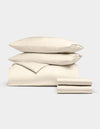 The Cozy Earth Bamboo Bedding Deluxe Bundle, featuring a cream comforter, two pillows, and two stacked sheets, is neatly folded and displayed against a plain light gray background.|Color:Creme