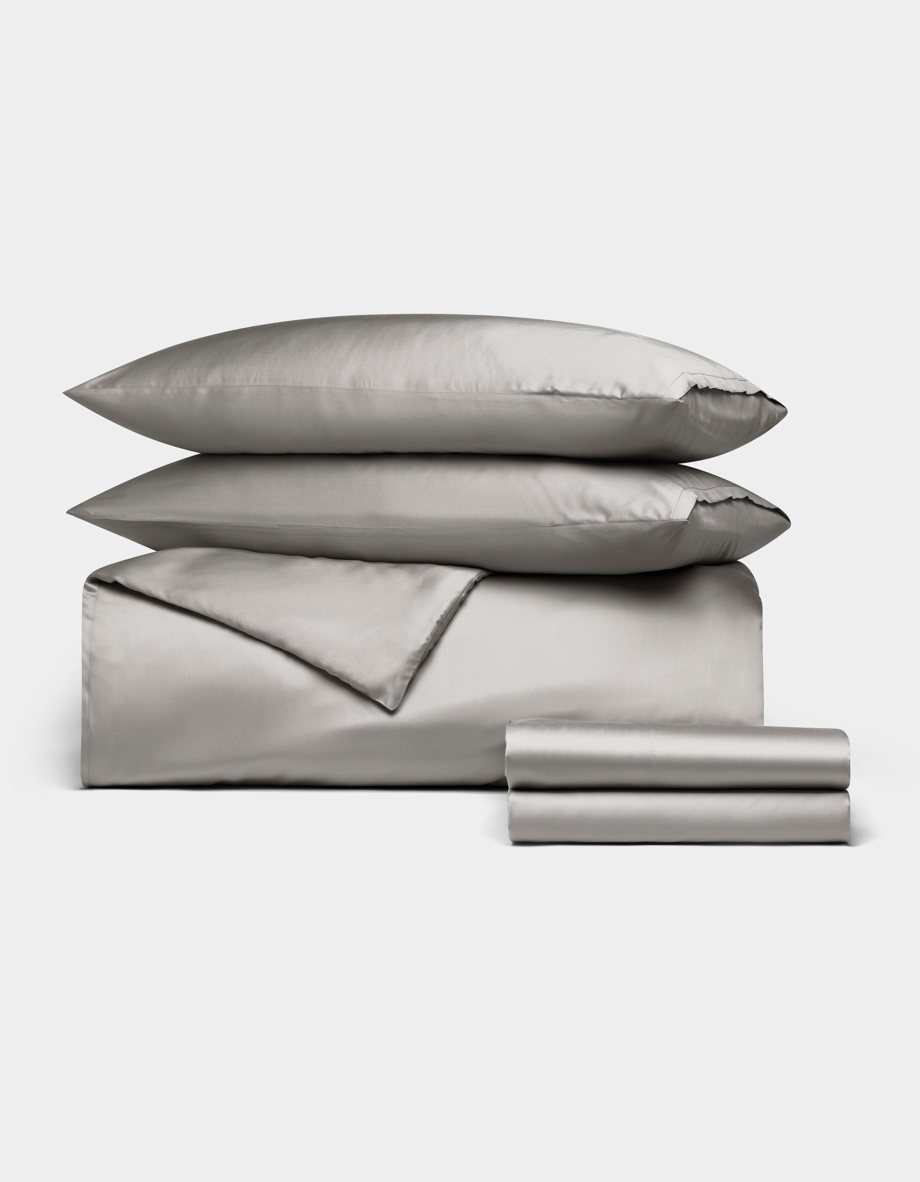 The Cozy Earth Bamboo Bedding Deluxe Bundle features light gray satin bedding with two pillows, a folded duvet, and stacked sheets, all neatly arranged on a white background.