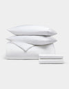The Bamboo Bedding Deluxe Bundle by Cozy Earth features a neatly stacked set of white bedding—two pillows, a comforter, and a folded sheet—displayed against a plain light gray background.|Color:White