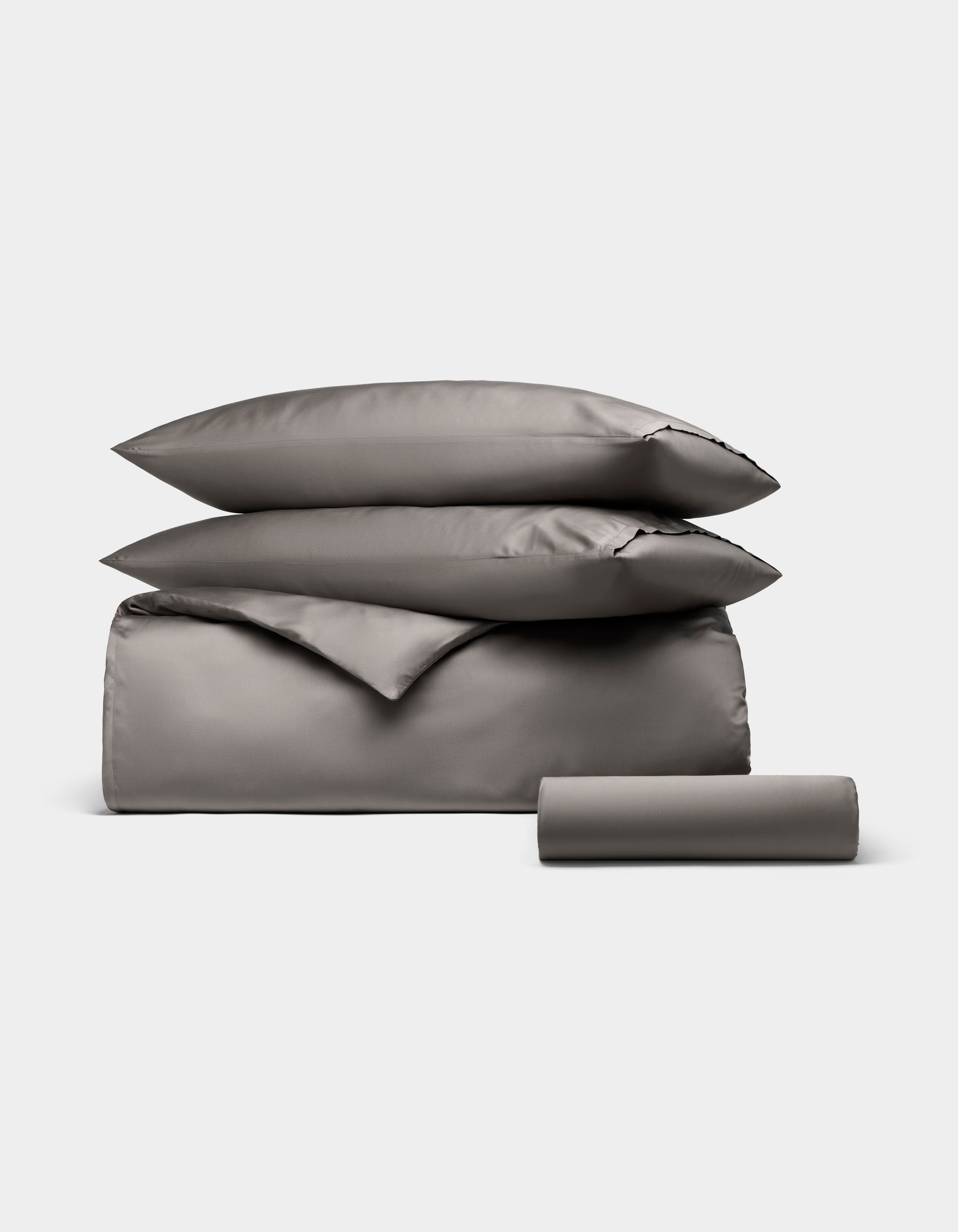 The Cozy Earth Bamboo Bedding Essential Bundle features a neatly stacked set of gray bedding—two pillows, a folded duvet, and a bolster pillow—displayed on a plain light background.|Color:Charcoal
