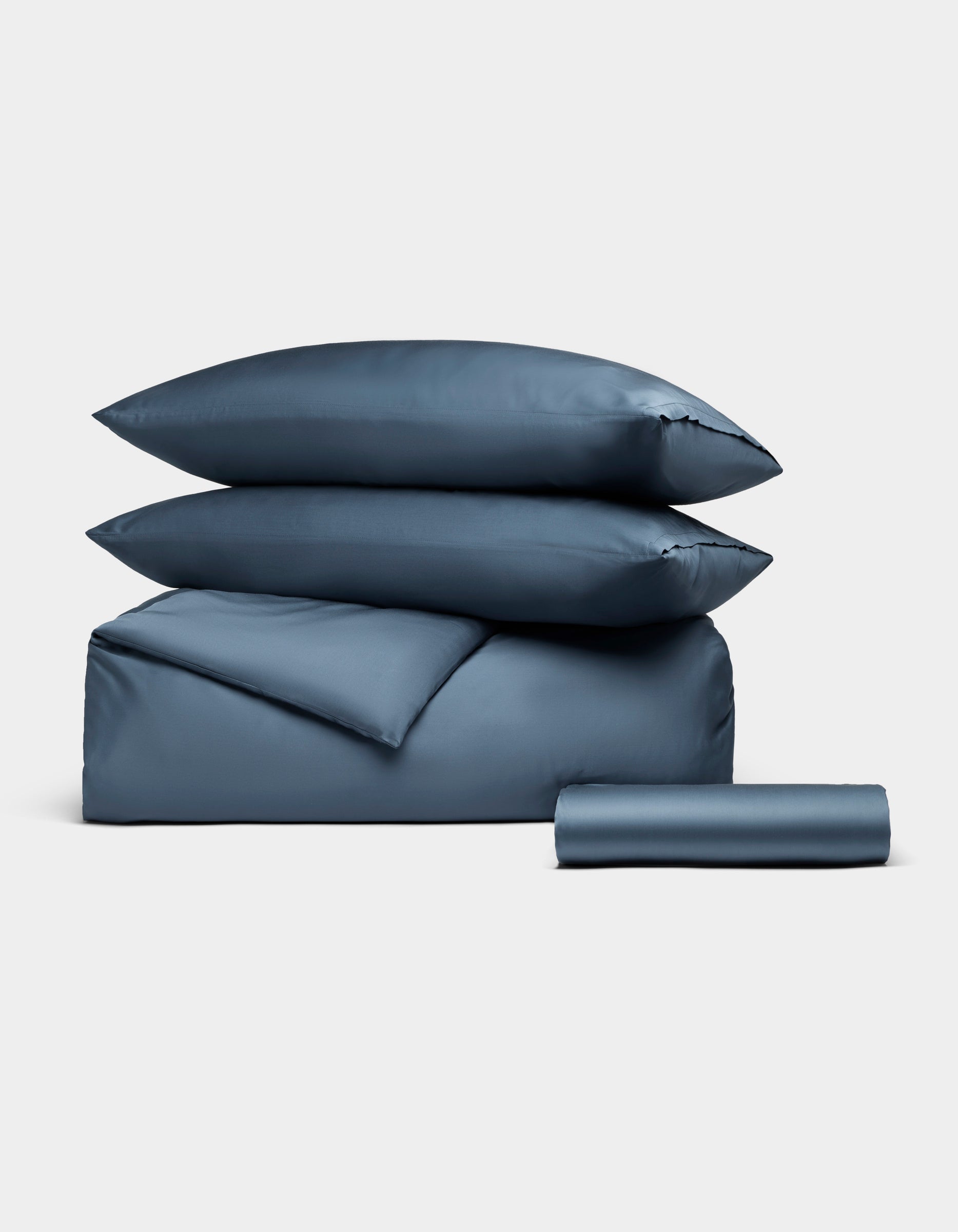 The Bamboo Bedding Essential Bundle by Cozy Earth features two pillows, a folded comforter, and a rolled bedsheet in blue, all neatly arranged against a plain white background.