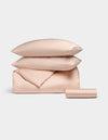 The Bamboo Bedding Essential Bundle by Cozy Earth, featuring light pink satin pillows, a folded duvet, and a rolled sheet, is neatly arranged against a light grey background.|Color:Peony