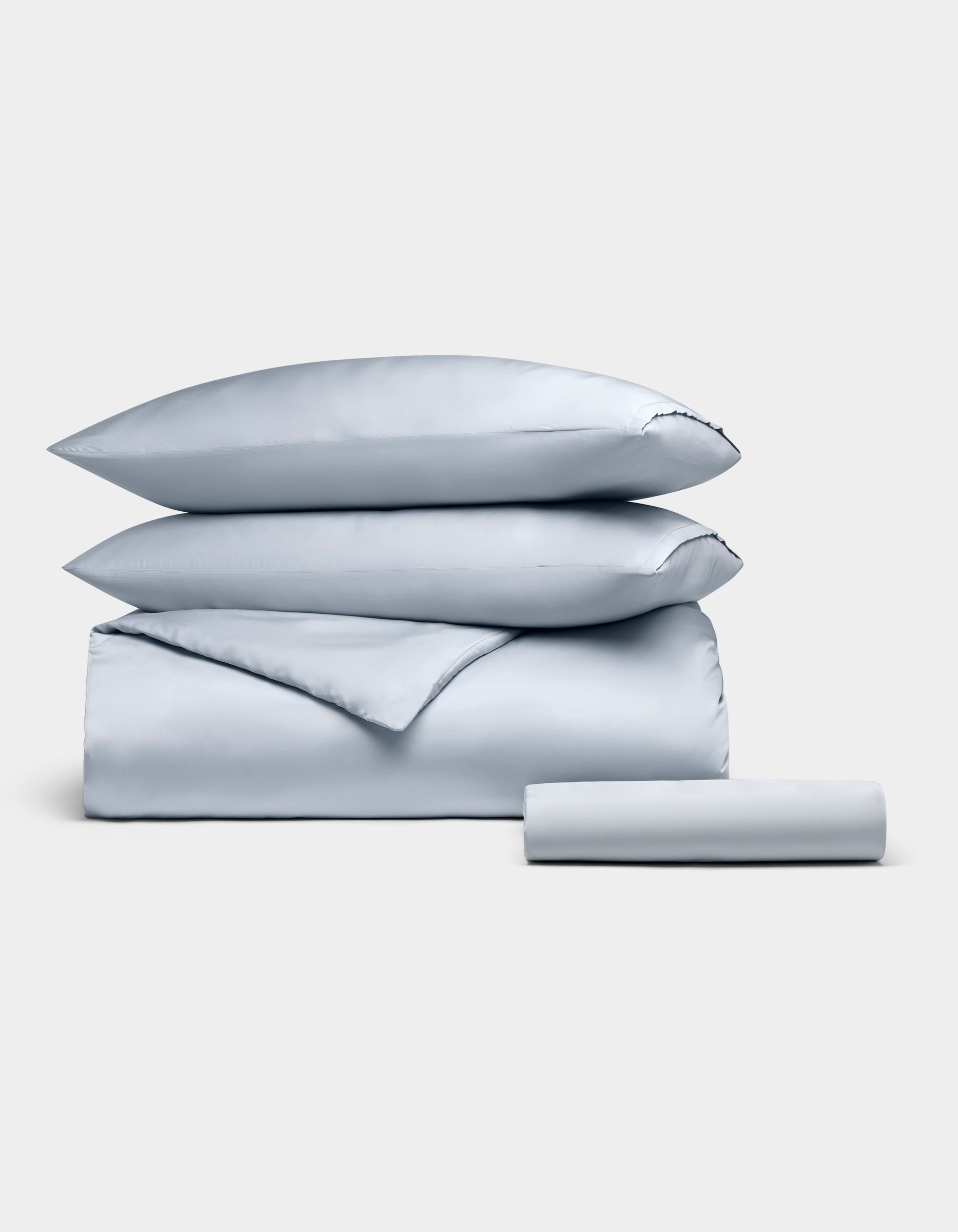 The Cozy Earth Bamboo Bedding Essential Bundle, featuring two pillows, a folded duvet, and a rolled sheet in light gray, is neatly stacked and displayed against a plain white background.|Color:Shore