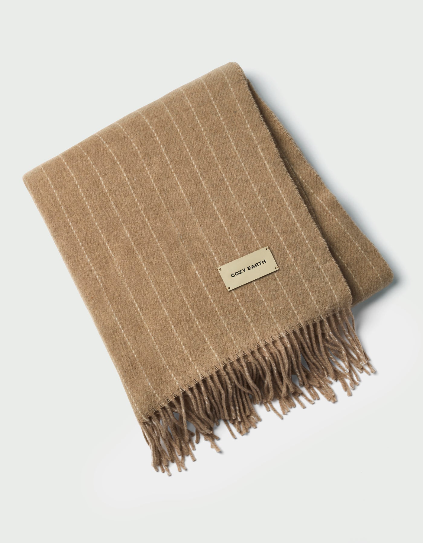 The Florence Throw by Cozy Earth is a tan, folded blanket with white pinstripes, fringed edges, and a rectangular "COZY EARTH" label on top, shown against a white background.|Color:Brandy