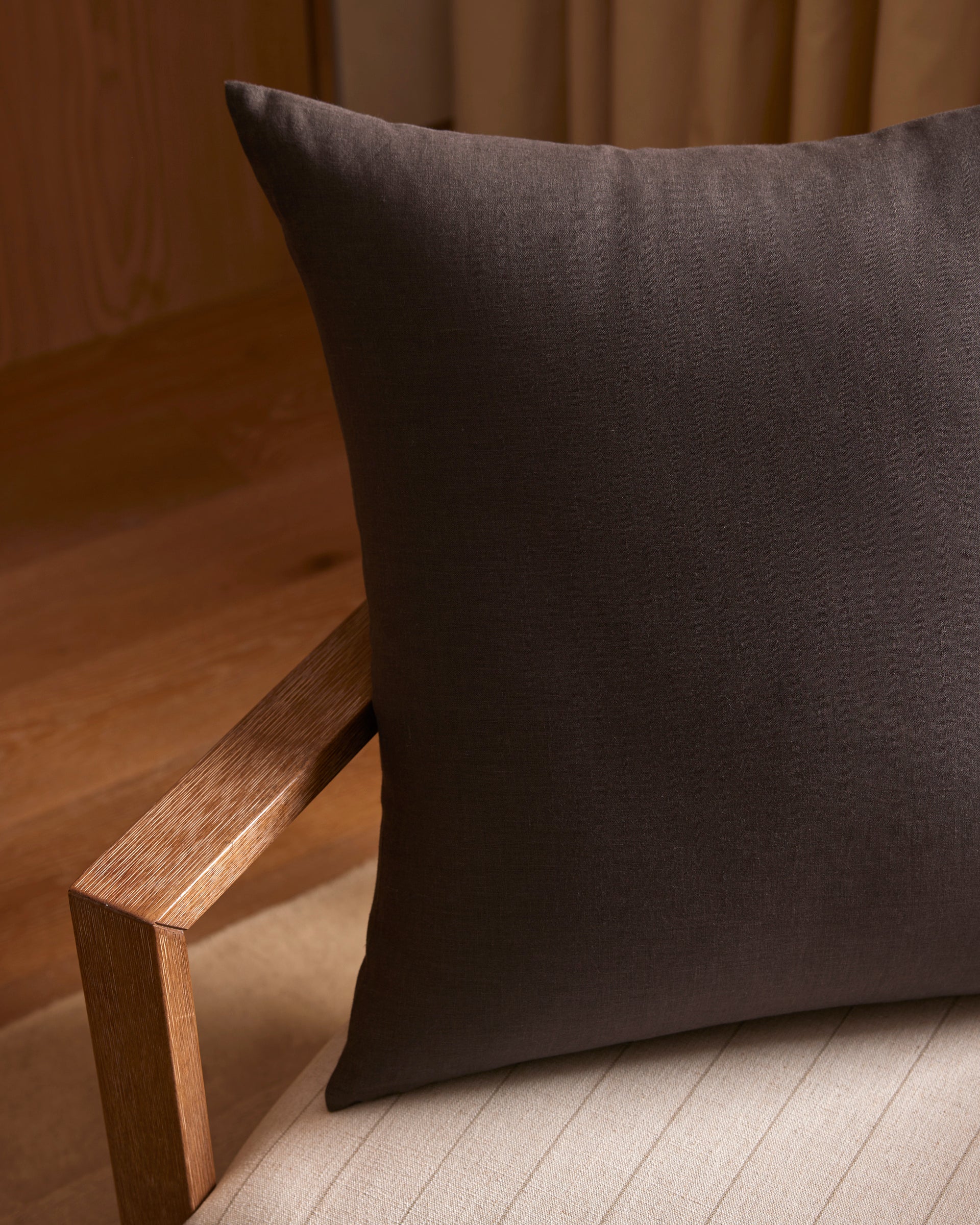 A close-up of the Cozy Earth Linen Throw Pillow in dark brown, placed on a light cushioned chair with a wooden armrest, set in a softly lit room featuring wooden flooring and beige curtains.|Color:Coal|Size:24" x 24"