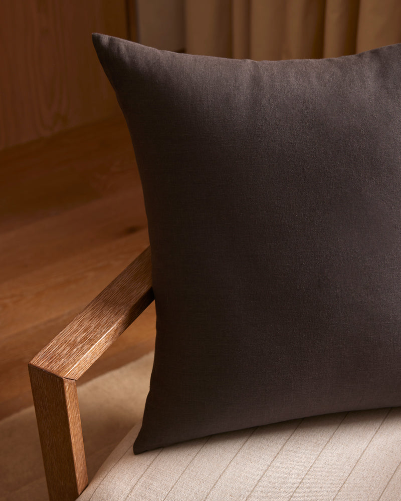 A Cozy Earth Linen Throw Pillow in dark brown rests on a light cushioned wooden chair, with wooden flooring and beige curtains in the background.