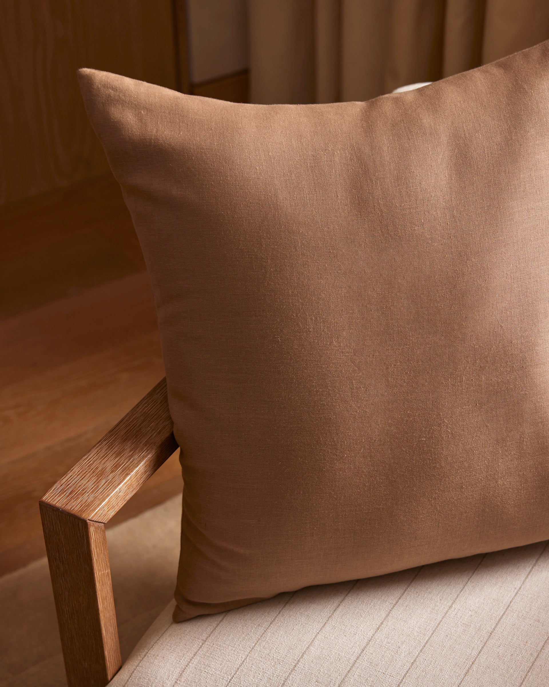 A Cozy Earth Linen Throw Pillow sits on a light beige upholstered chair with a wooden armrest, in a softly lit room featuring wooden flooring and neutral curtains.