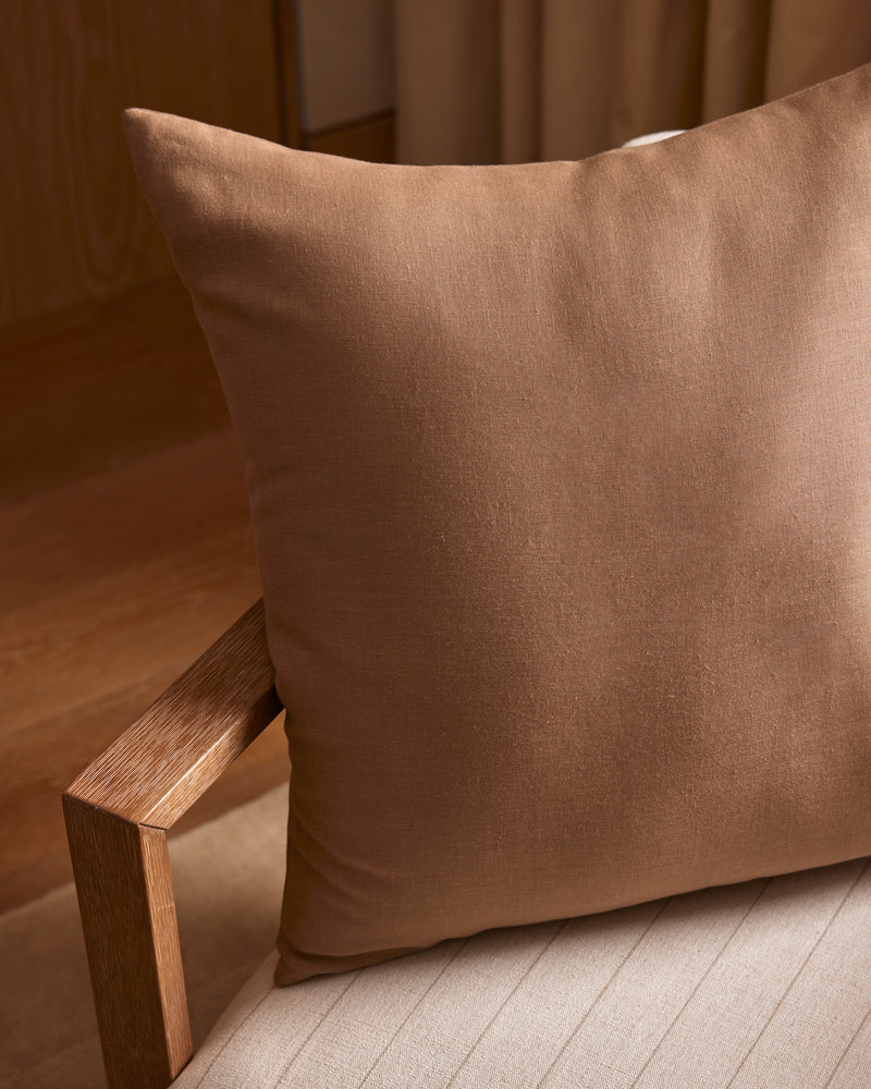 A Linen Throw Pillow by Cozy Earth sits on a beige, striped cushioned chair with a wooden armrest, in a warmly lit room featuring wooden floors and neutral-colored walls.