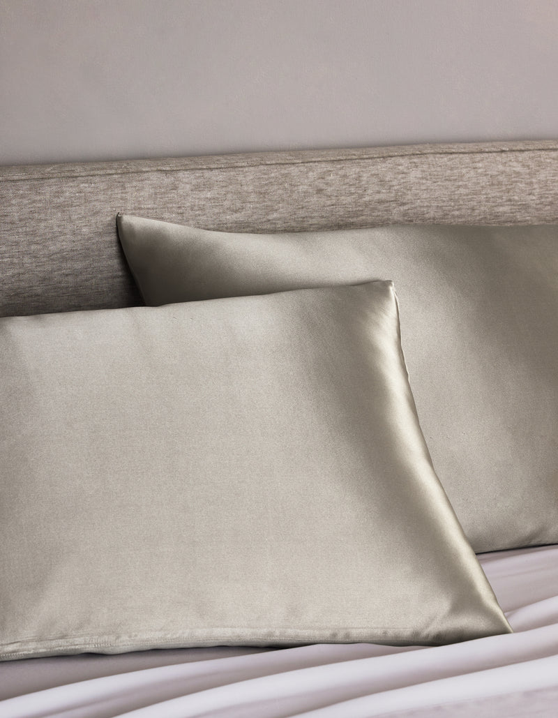 Two Cozy Earth Silk Pillowcases in light gray are arranged on a bed with a light beige upholstered headboard, adding a smooth, shiny, and luxurious touch to the bedding.