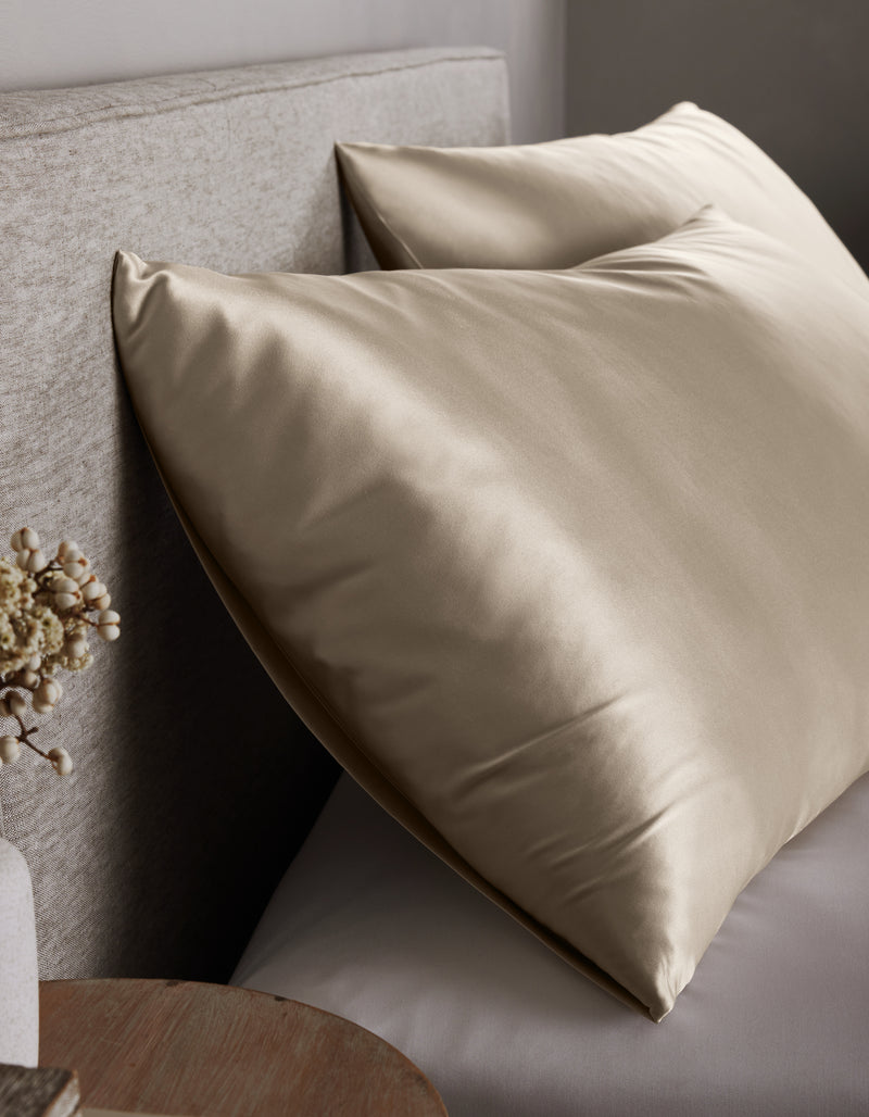 Two large, shiny Cozy Earth Silk Pillowcases in champagne rest against a light upholstered headboard on the bed, with a small round wooden nightstand and a vase of dried flowers nearby.