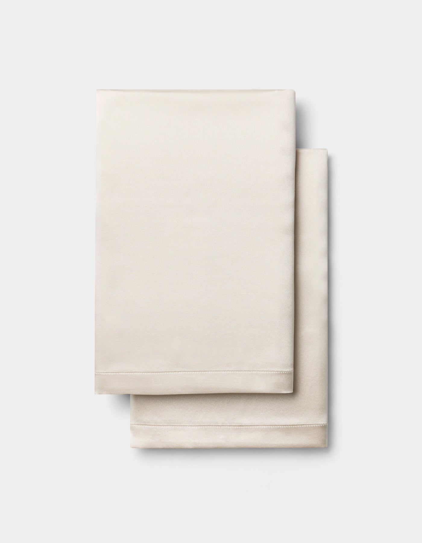 Two neatly folded, light beige Silk Pillowcases by Cozy Earth are stacked on a plain, light gray background.|Color:Oat
