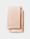 Two neatly folded Cozy Earth Silk Pillowcases in light peach are stacked on a light gray background.|Color:Peony