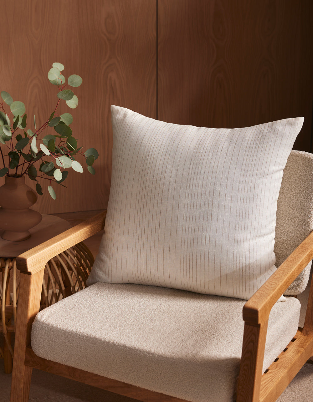 A wooden armchair with a cream cushion and the Cozy Earth Striped Throw Pillow sits next to a small wicker table holding a brown vase with eucalyptus branches, set against a wooden wall.