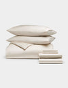 The Cozy Earth Bamboo Bedding Ultimate Bundle, featuring ivory-colored bed linens with two pillows, a folded comforter, and stacked sheets, is displayed against a plain white background.|Color:Oat