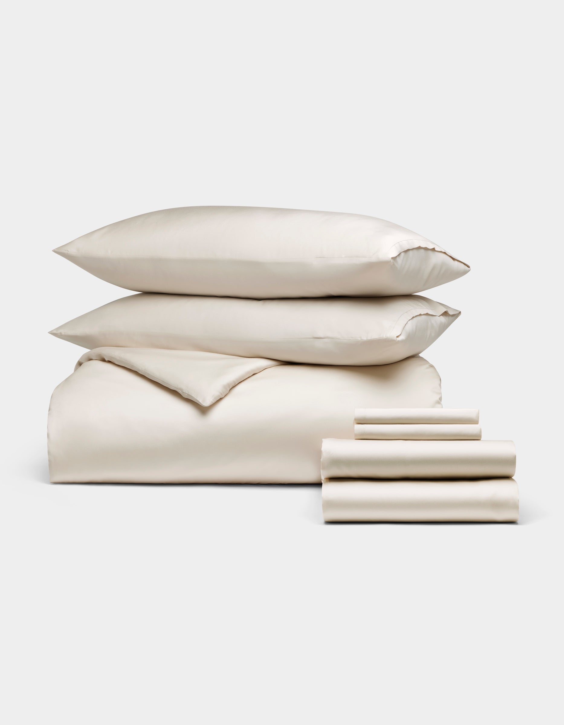 The Cozy Earth Bamboo Bedding Ultimate Bundle, featuring ivory-colored bed linens with two pillows, a folded comforter, and stacked sheets, is displayed against a plain white background.