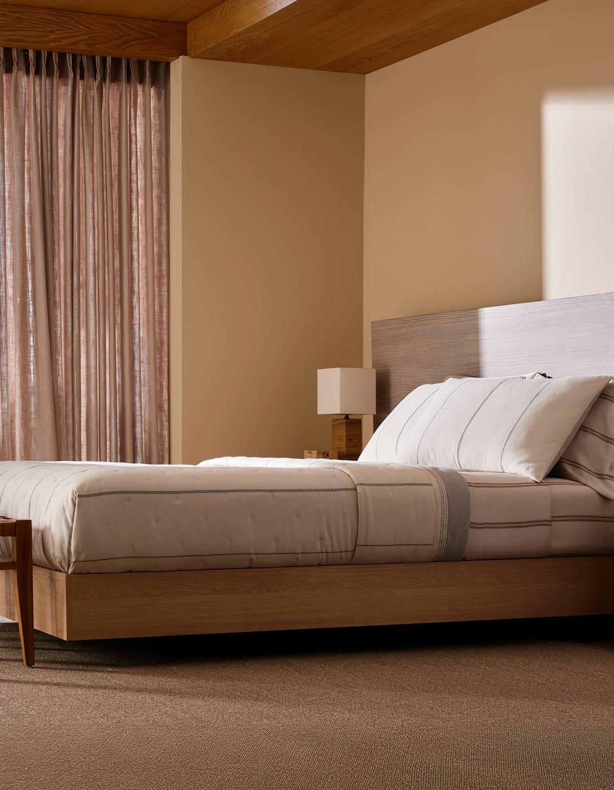A modern bedroom features Cozy Earth’s Baja Quilt Bundle on a wooden platform bed, paired with a matching nightstand and lamp. Sheer curtains let in soft natural light, fostering a calm, minimalistic vibe.|Color:Coal Stripe/Striped Alabaster