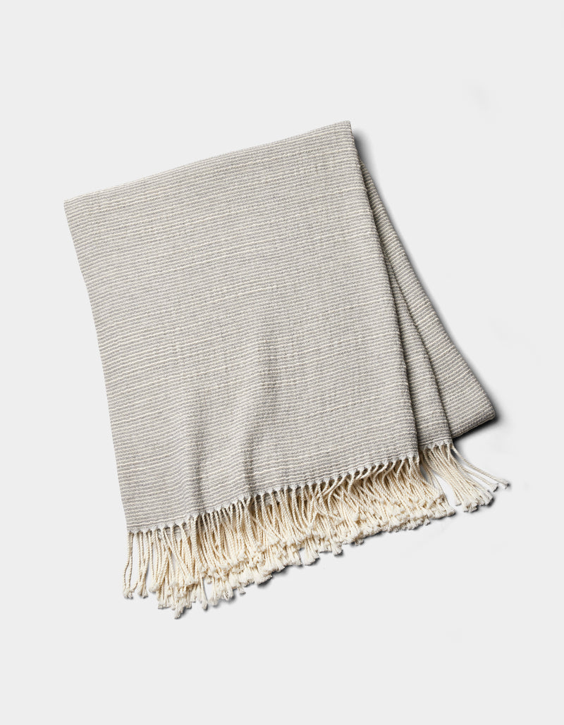 The Cozy Earth Baja Tassel Throw is a folded light gray woven blanket with subtle stripes and cream fringe on the ends, shown on a white background.