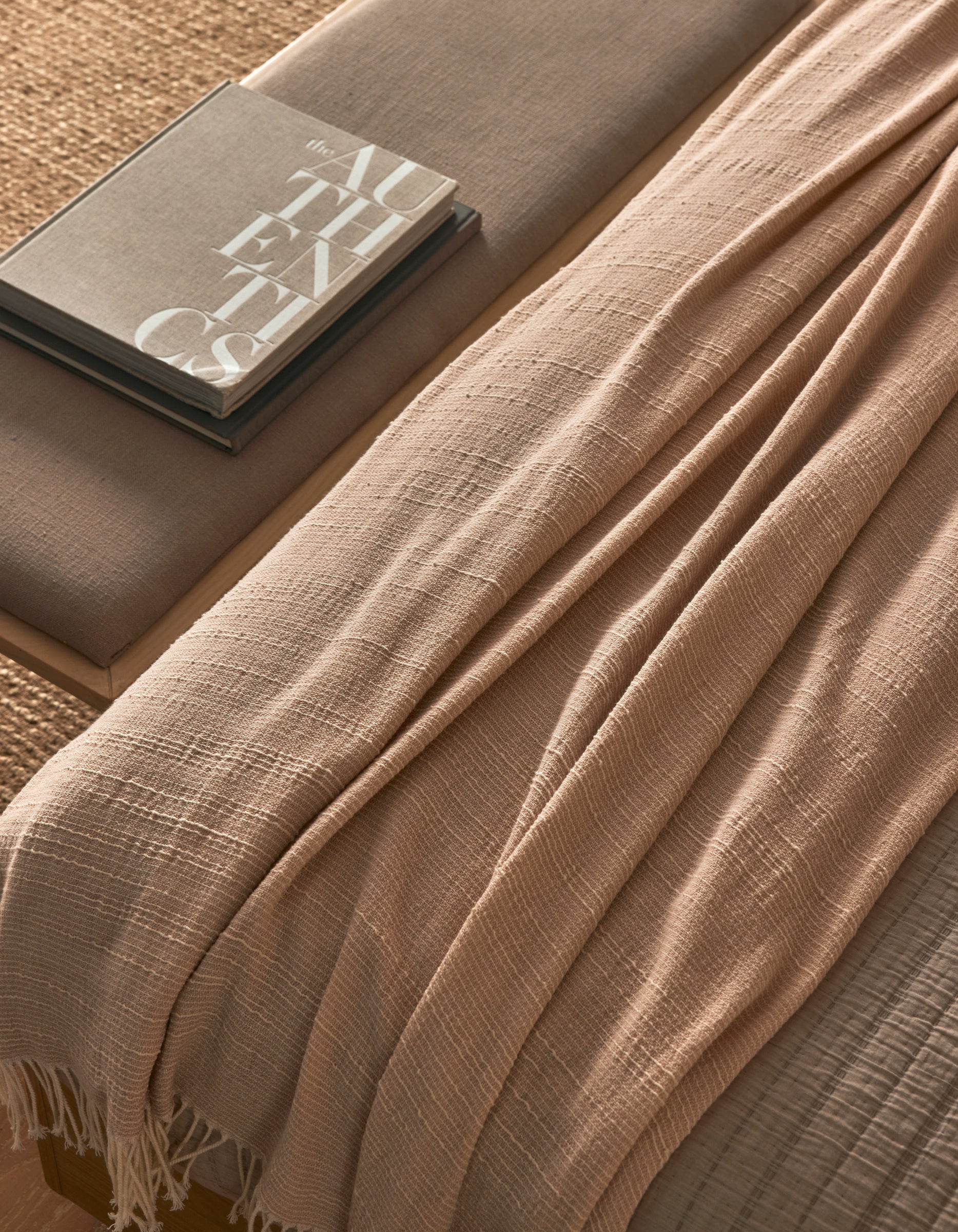 The Baja Tassel Throw by Cozy Earth, in beige with a textured finish, is draped over a neutral-toned bench beside two stacked books (one titled "AUTHENTICS"), creating a cozy, minimalistic look.