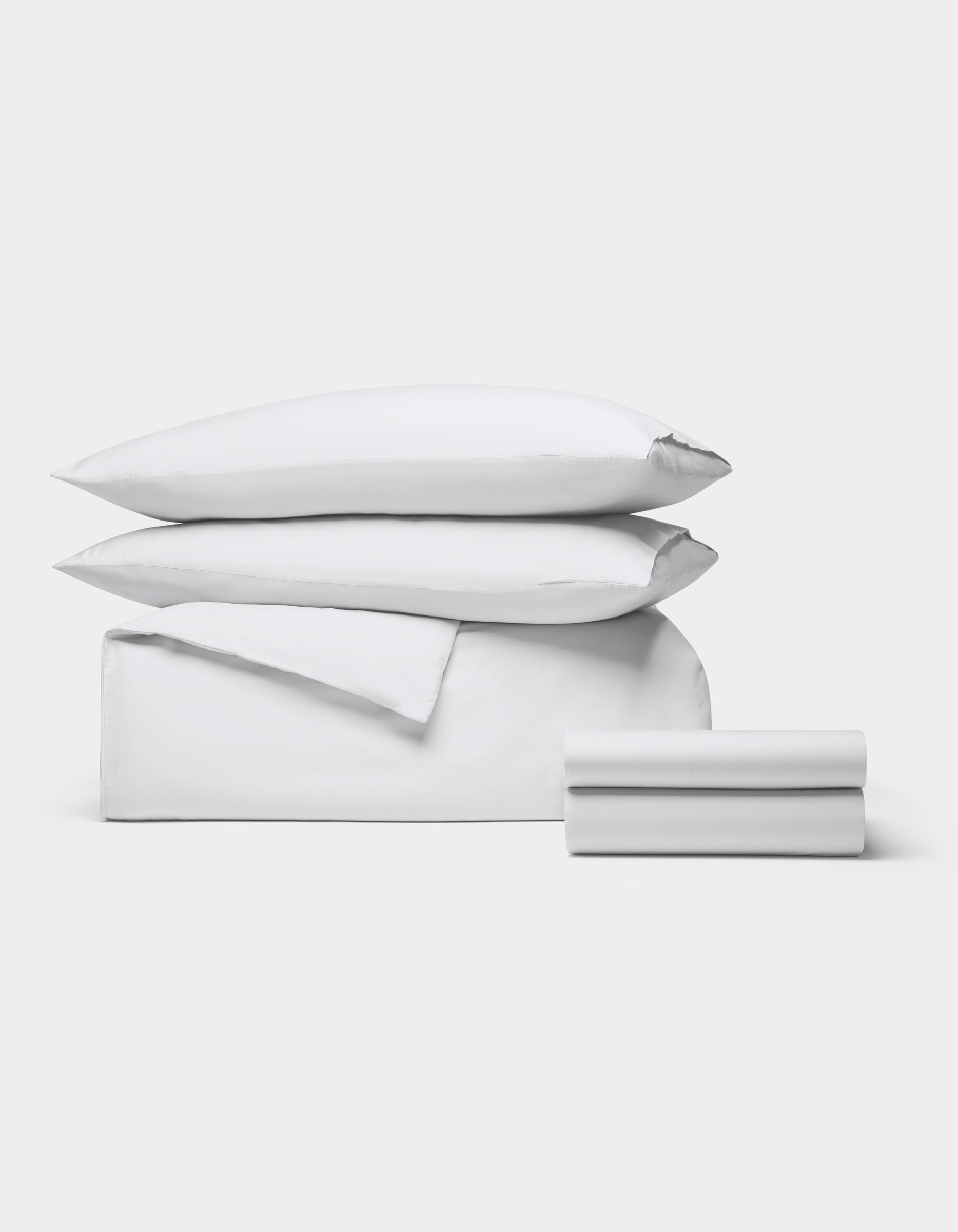 The Cozy Earth Soft-Wash Cotton Bedding Deluxe Bundle, featuring two pillows, a duvet cover, and two sheets in white, is neatly stacked against a plain light gray background.
