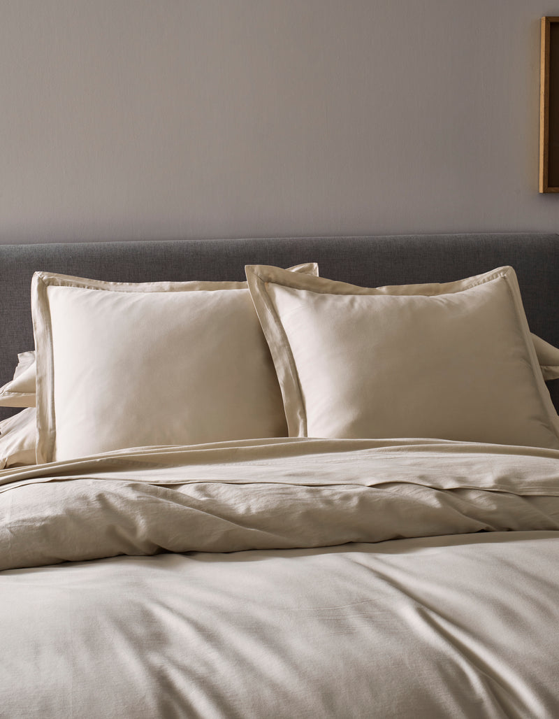 Two Soft-Wash Cotton Shams by Cozy Earth in beige rest against a gray upholstered headboard, paired with a coordinated beige comforter and sheets, creating a neat, minimal look in this modern bedroom.