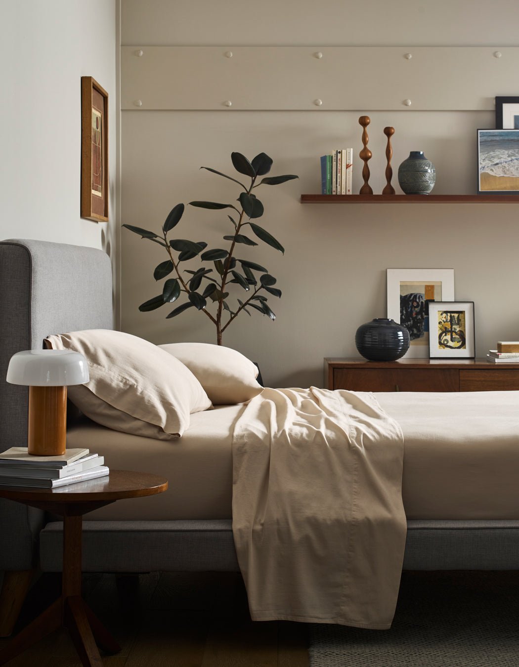 A neatly made bed with Cozy Earth Soft-Wash Cotton Pillowcases sits beside a wooden nightstand with a white lamp. A large green plant, artwork, and decor items decorate the dresser and a wooden shelf on the wall.