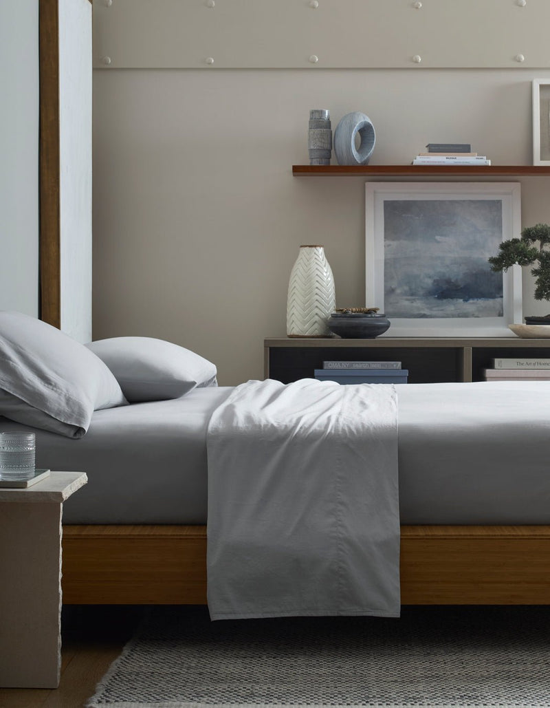 A neatly made bed with Cozy Earth Soft-Wash Cotton Flat Sheets sits by a nightstand holding a glass of water. Shelves behind display books, vases, and framed art, creating a serene and modern bedroom atmosphere.