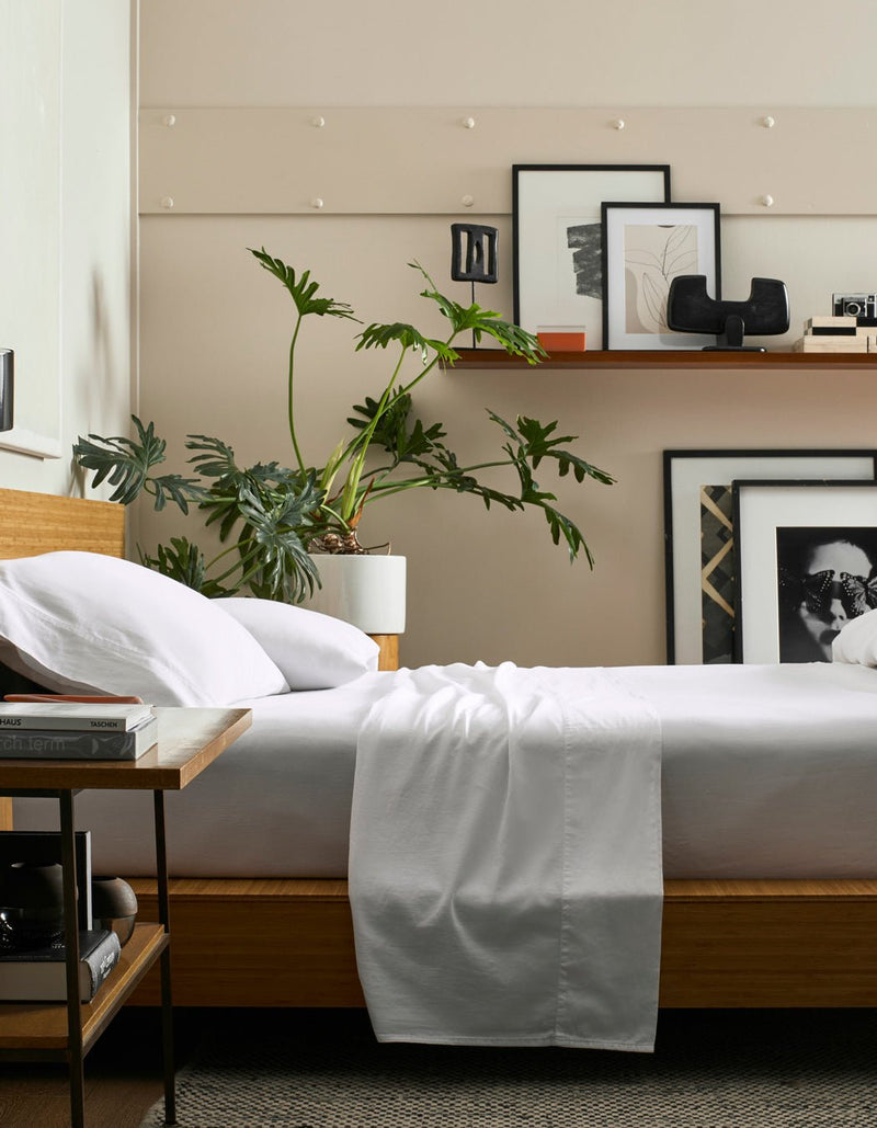 A modern bedroom featuring a Cozy Earth Soft-Wash Cotton Fitted Sheet on a wooden bed, with white bedding, a leafy plant, floating shelf holding framed art above the bed, and a side table with books.