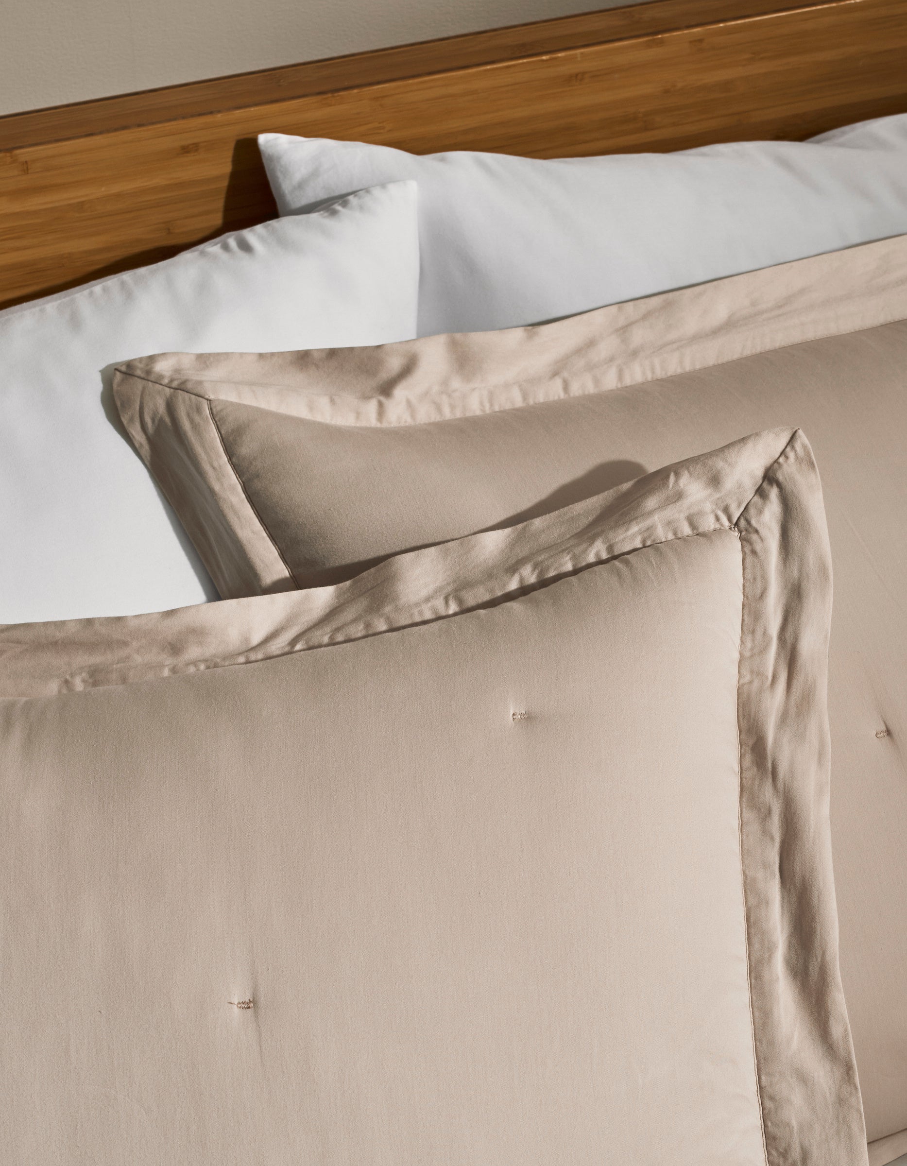 Two Cozy Earth Soft-Wash Cotton Quilted Shams in beige rest against a white pillow on a wooden headboard bed, creating a clean and cozy bedroom setting.
